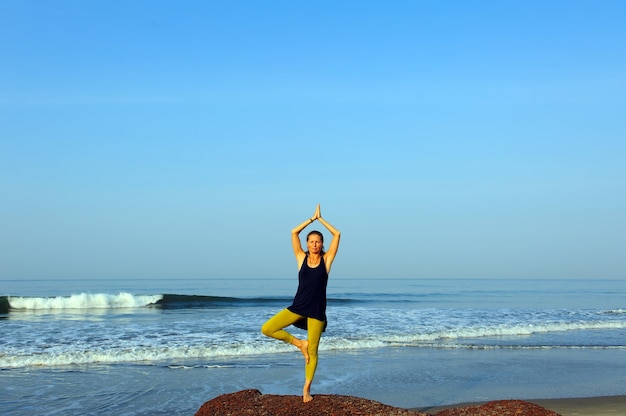 Hermosa joven practicando yoga y ejercicios de estiramiento en la playa del océano de verano