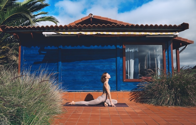 Hermosa joven practicando yoga en el suelo cerca de la casa de madera azul