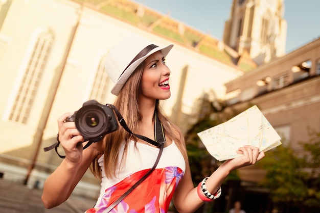 Hermosa joven preocupada de vacaciones caminando por la ciudad y sosteniendo un mapa de la ciudad y una cámara digital.