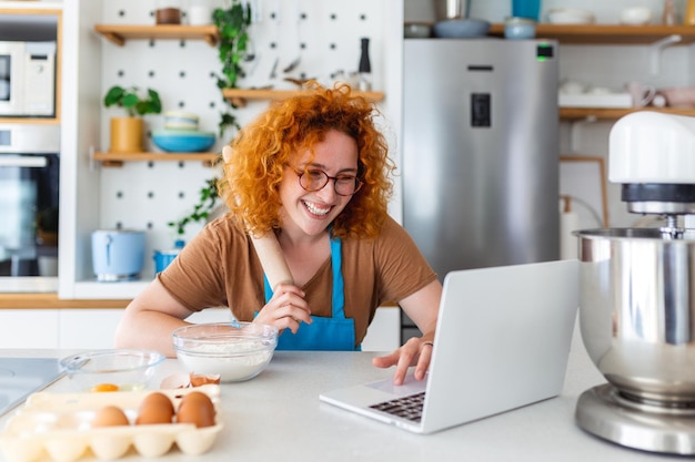 Hermosa joven profesional feliz está blogueando para su canal de cocina sobre una vida saludable en la cocina de su casa y mirando a la cámara en una computadora portátil