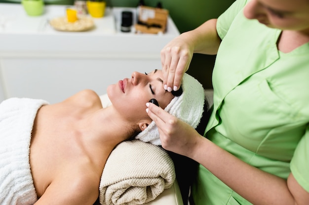 Hermosa joven recibiendo masaje facial con piedras calientes en el spa de belleza. Terapia Lastone.