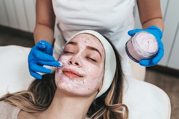 Hermosa joven recibiendo mascarilla facial con efectos rejuvenecedores en el salón de belleza spa.