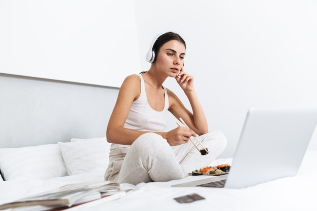 Hermosa joven relajándose en la cama en casa, escuchando música con auriculares, comiendo sushi de un plato