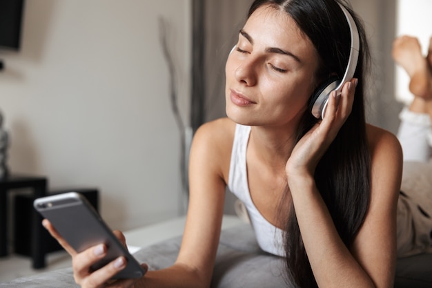 Hermosa joven relajándose en un sofá en casa, escuchando música con auriculares, sosteniendo el teléfono móvil