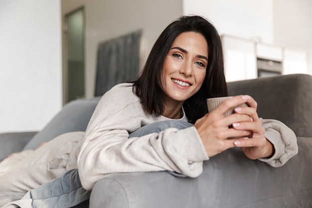 Hermosa joven relajándose en un sofá en casa, sosteniendo la taza