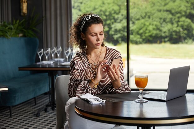 Hermosa joven rizada sentada en un café bebiendo jugo de naranja y esperando a su amiga