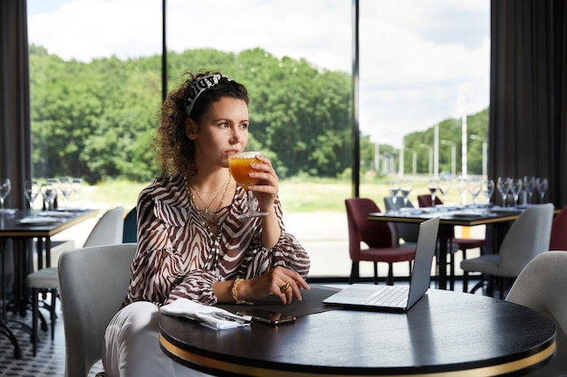 Hermosa joven rizada sentada en un café bebiendo jugo de naranja y esperando a su amiga