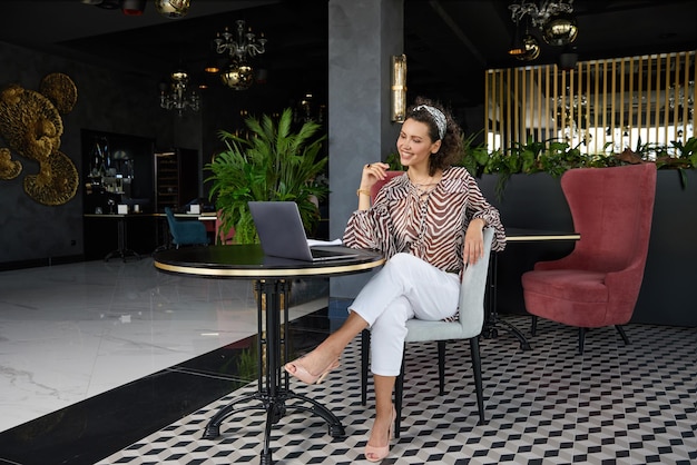 Hermosa joven rizada sentada en un café bebiendo jugo de naranja y esperando a su amiga