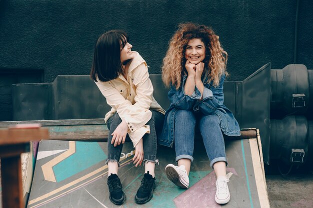 Hermosa joven rizada vestida con jeans mirando a la cámara riendo mientras su mejor amiga la mira sonriendo al aire libre.