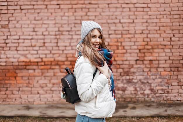 Hermosa joven en ropa de abrigo con bolsa posando junto a la pared de ladrillo