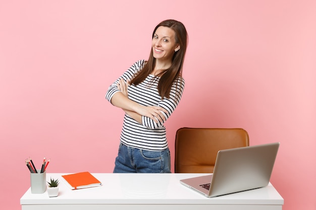 Hermosa joven en ropa casual de trabajo, de pie junto a un escritorio blanco con un portátil pc contemporáneo