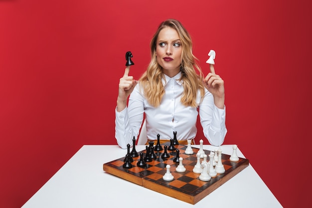 Hermosa joven rubia con camisa blanca sentado en la mesa, jugando al ajedrez aislado sobre fondo rojo.