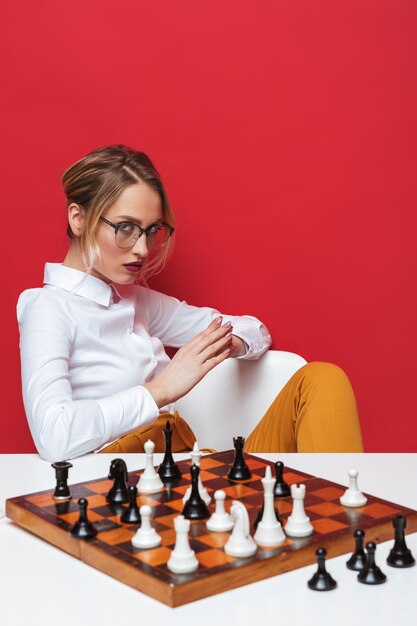 Hermosa joven rubia con camisa blanca sentado en la mesa, jugando al ajedrez aislado sobre fondo rojo.