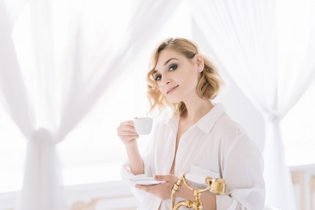 Hermosa joven rubia con una camisa ligera en una habitación luminosa disfruta de una taza de café por la mañana antes de ir a trabajar. Chica en el baño con ropa sincera.