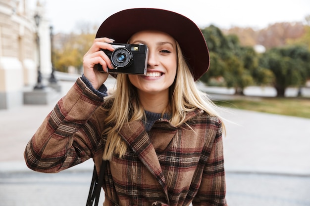 Hermosa joven rubia elegante con un abrigo caminando al aire libre, tomando fotografías con cámara de fotos