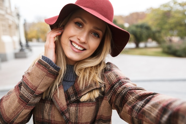 Hermosa joven rubia elegante con un abrigo caminando al aire libre, tomando un selfie con teléfono móvil