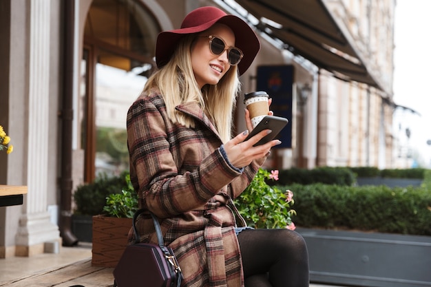 Hermosa joven rubia elegante vistiendo un abrigo relajante al aire libre, mediante teléfono móvil