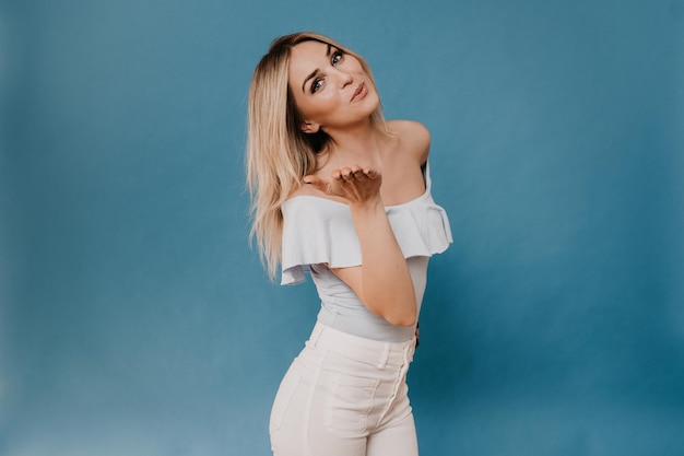 Hermosa joven rubia encantadora vestida con jeans blancos y blusa ligera dando un beso de aire sobre b