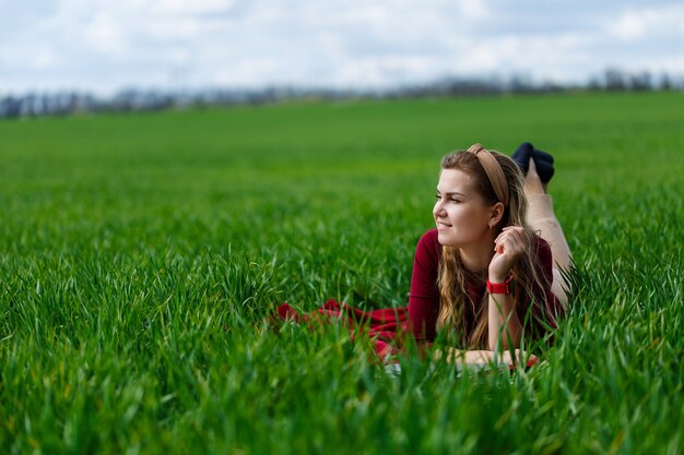 Hermosa joven rubia está acostada sobre la hierba verde en el parque con una computadora portátil y trabajando. Cielo azul con nubes. La niña sonríe y disfruta de un buen día. Trabaja en la naturaleza en un día soleado.