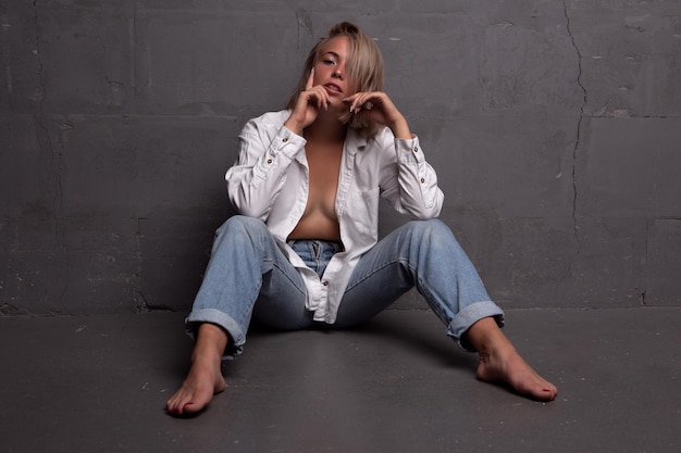 Hermosa joven rubia está sentada en el suelo con jeans y una camisa desabrochada