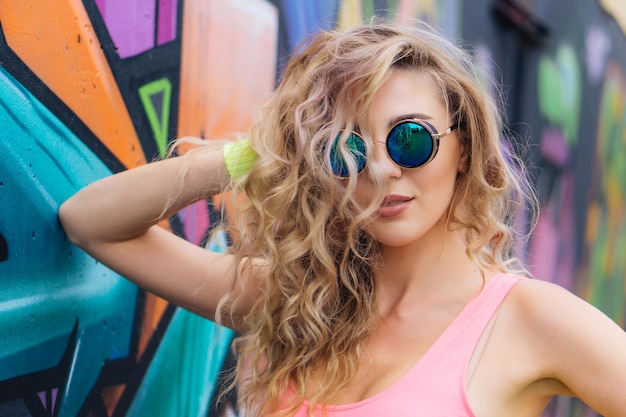 Hermosa joven rubia con gafas de sol junto a la pared con graffiti