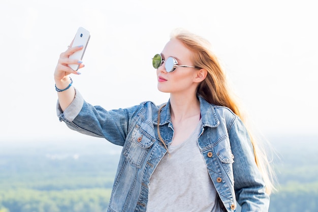 Hermosa joven rubia con gafas de sol se toma una selfie contra la naturaleza del paisaje