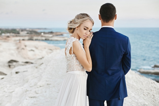 Hermosa joven rubia modelo en vestido de encaje blanco de moda apoyarse en el hombre guapo en el elegante traje azul y posando en la roca blanca en la costa del mar Adriático