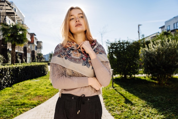 Hermosa joven rubia con un pañuelo sobre los hombros camina, disfruta del cálido clima primaveral y del sol