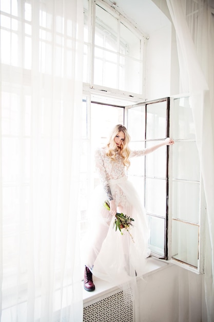 Hermosa joven rubia prometida con maquillaje de evento de peinado nupcial y flores abriendo la ventana