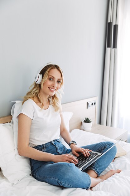 Hermosa joven rubia relajándose en la cama en casa, escuchando música con auriculares, trabajando en equipo portátil