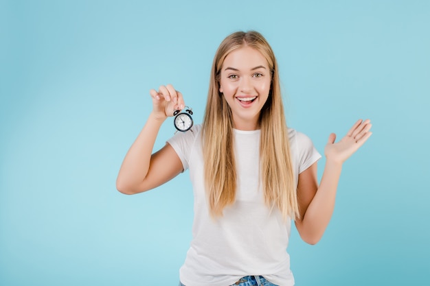 Hermosa joven rubia con reloj despertador mostrando tiempo aislado sobre azul