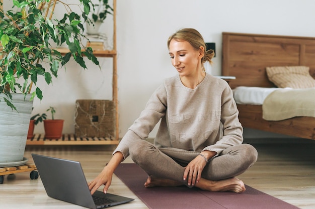 Hermosa joven rubia con ropa deportiva cómoda practica yoga en línea en un dormitorio acogedor en casa