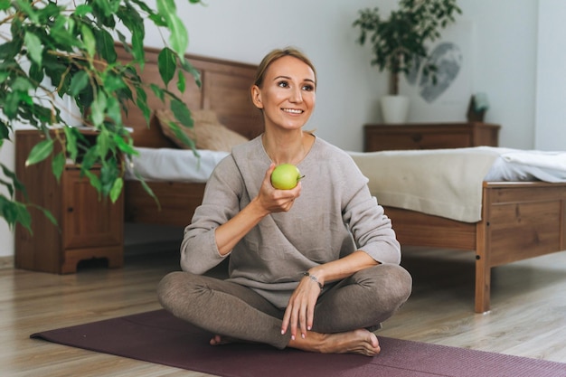 Hermosa joven rubia en ropa deportiva práctica yoga mantenga manzana verde en la mano en casa