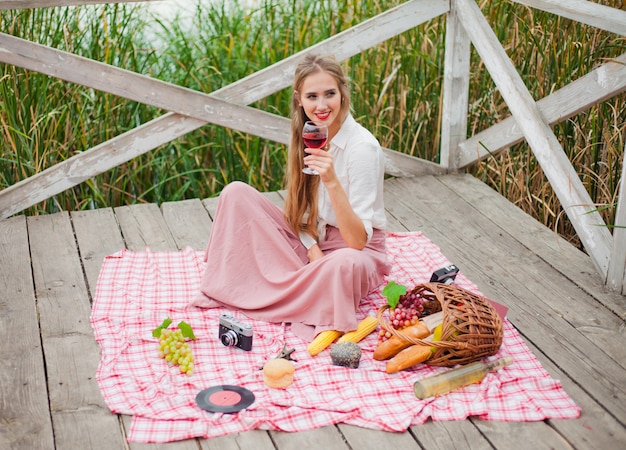 Hermosa joven rubia en ropa retro vintage con picnic en el muelle de madera solo. Picnic estilo francés al aire libre