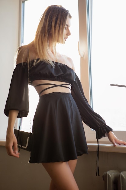 Una hermosa joven rubia sexy con un vestido corto negro de seda está de pie junto a la ventana