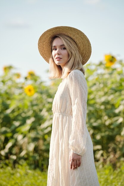 Hermosa joven rubia con sombrero de paja y vestido romántico blanco de pie delante de la cámara contra el campo de girasol en un día soleado