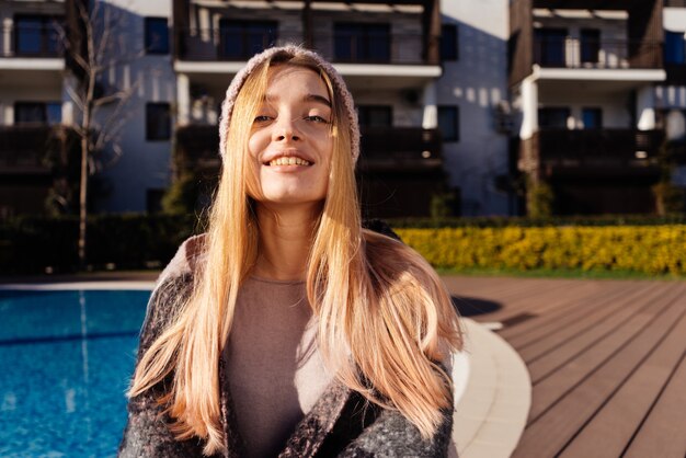 Hermosa joven rubia con sombrero se sienta junto a la piscina al aire libre, bajo el sol, mira a la cámara y sonríe