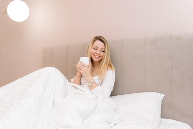 Hermosa joven rubia sonriente con café en una cama blanca buenos días