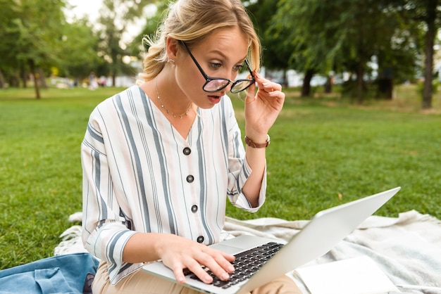 Hermosa joven rubia sorprendida relajándose en un césped en el parque, trabajando en la computadora portátil mientras está sentado en una manta