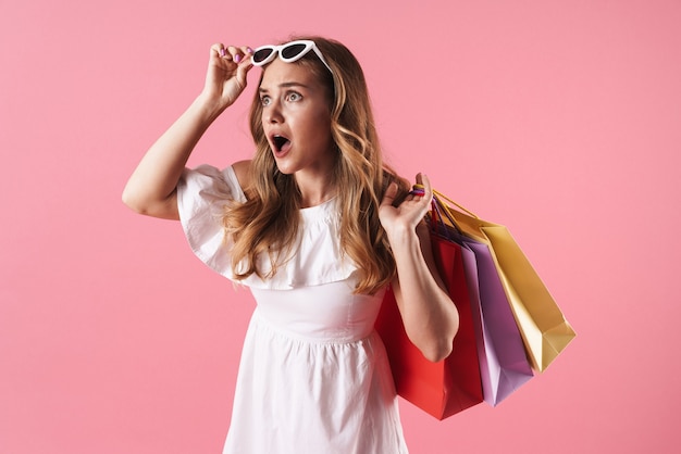 Hermosa joven rubia sorprendida con vestido de verano que se encuentran aisladas sobre la pared rosa, llevando bolsas de la compra.