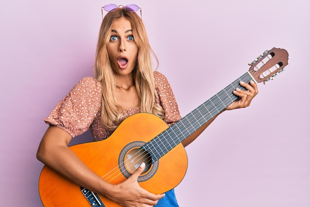 Hermosa joven rubia tocando guitarra clásica asustada y conmocionada con sorpresa y expresión asombrada miedo y cara emocionada