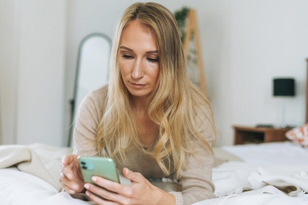 Hermosa joven rubia usando el móvil en la cama en una habitación luminosa en el hogar