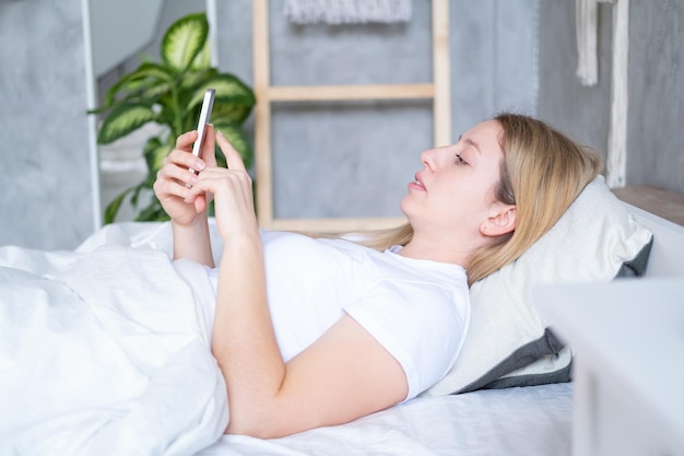 Hermosa joven rubia usando teléfono móvil acostada en la cama en un interior moderno