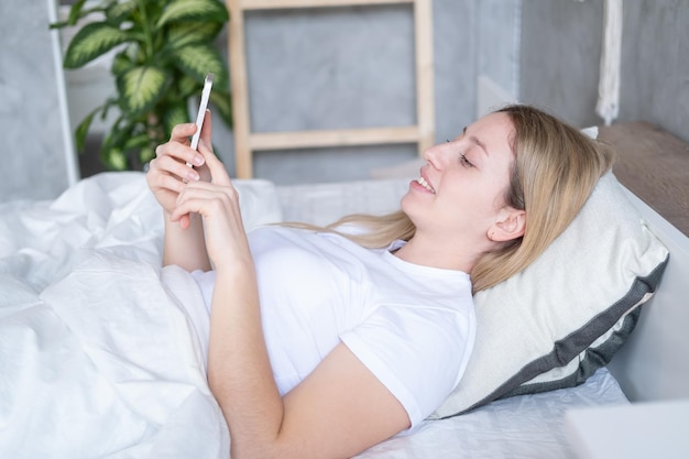 Hermosa joven rubia usando teléfono móvil acostada en la cama en un interior moderno