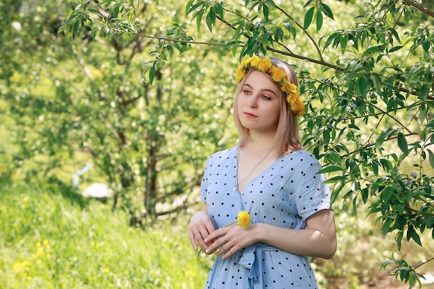 Hermosa joven rubia en vestido azul claro y corona de diente de león en el jardín de verano