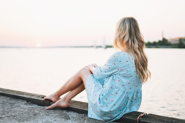 Hermosa joven rubia con vestido azul sentada en el muelle y mirando la puesta de sol