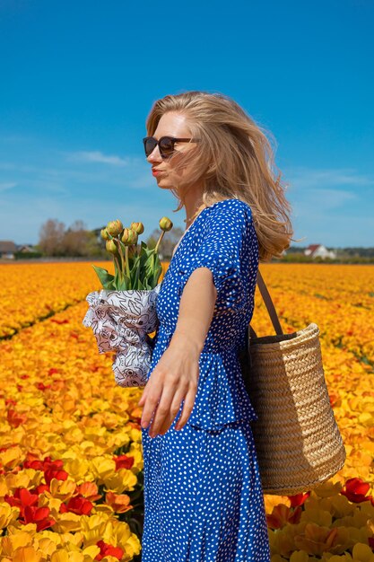 Hermosa joven rubia con un vestido azul sosteniendo una cesta de paja en un campo de tulipanes de flores amarillas y naranjas en un día soleado de verano contra un fondo de cielo azul claro Concepto de viaje por la naturaleza