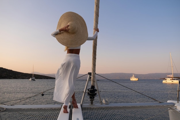 Hermosa joven rubia con un vestido de pie en la popa del yate en el mar o el océano al atardecer