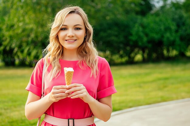 Y una hermosa joven rubia con un vestido rosa comiendo helado de vainilla en un cono de galleta en el parque de verano