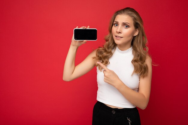 Hermosa joven rubia vistiendo camiseta blanca aislada sobre fondo rojo con espacio de copia sosteniendo